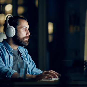 Man wearing white headphones and a denim shirt, working at a computer in a dimly lit room at night.